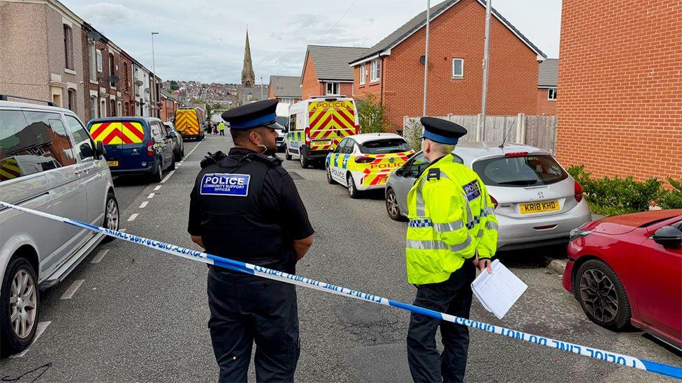 Police officers stand behind blue-and-white police tape on a residential street lined with terraced and new-build houses. Several marked police cars and emergency service vehicles are parked along the road, which has been cordoned off
