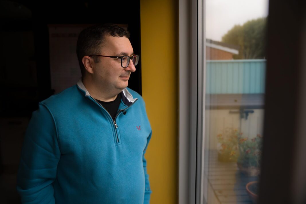 Thornton-Cleveleys resident Andrew Molloy looks out of the window in his home. He is wearing a blue jumper and glasses.