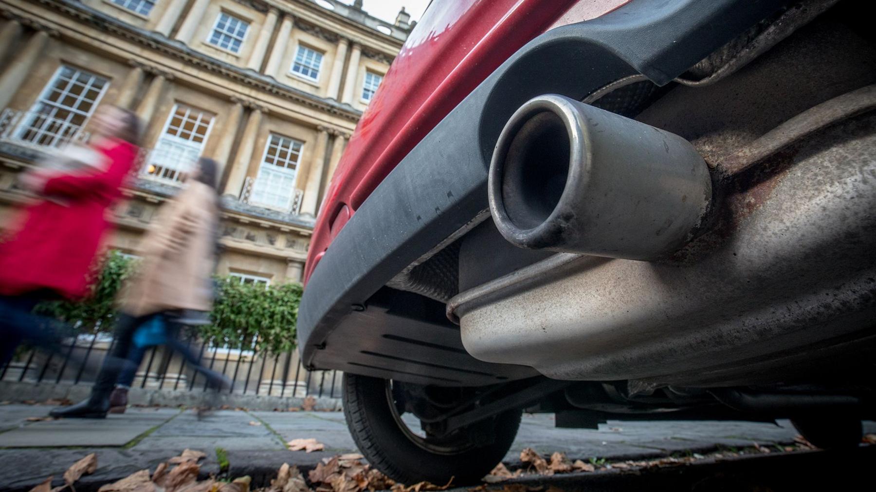 Close up of an exhaust pipe on the back of a red car on a street in the UK. There are Autum leaves on the ground  and two blurred poeple walking in the distance