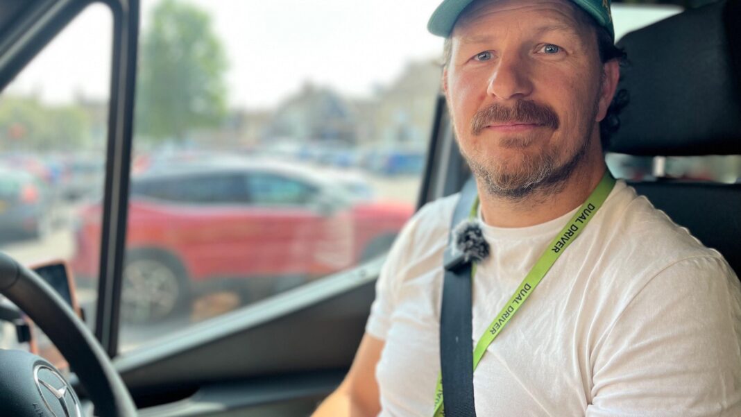 Countryside buses resurging in England after years of decline – check your local figures James smiles at the camera. He is sitting in the driver's seat of a minibus and is wearing a white T-shirt and a cap. He has a light green lanyard around his neck and is wearing his seatbelt. Parked cars can be seen through the window of the minibus.