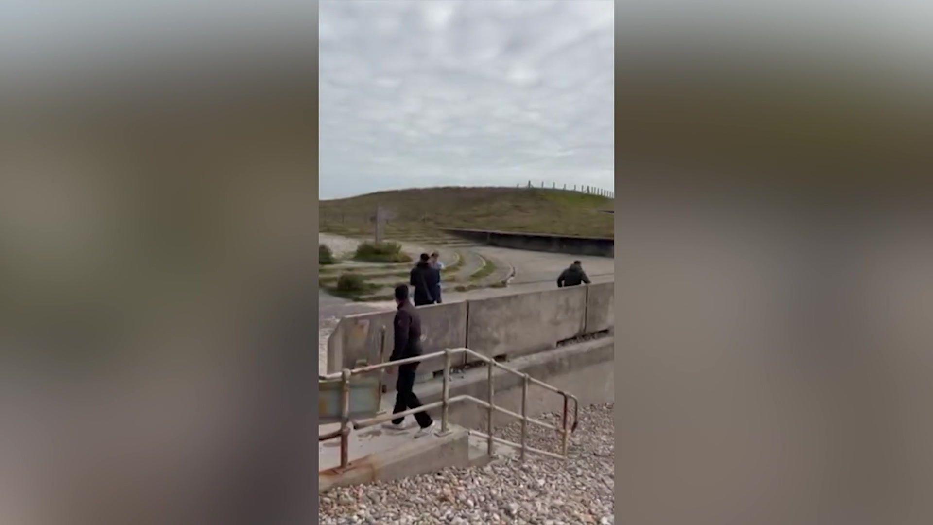 Vertical phone video shows several people walking along a concrete sea-defence structure beside a wide pebble beach. Metal handrails lead down steps in the foreground, while curving concrete channels and a low embankment stretch across the distance. A grassy hill topped with a fence runs along the horizon beneath a grey sky.