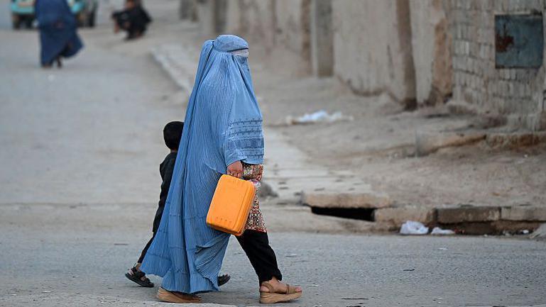 An Afghan burqa-clad woman walks along a street in the 7th district of Afghanistan's Kandahar, several people are behind her. She is walking next to a child. The street is dusty and beige.
