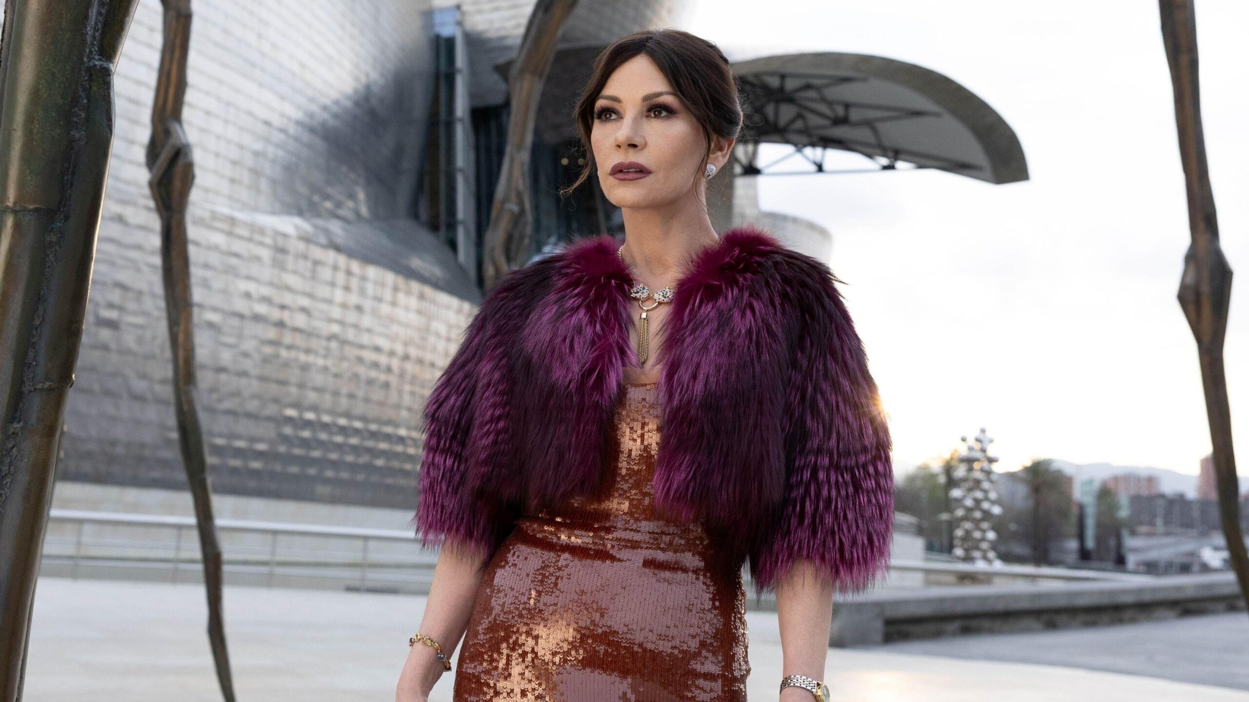 Catherine Zeta-Jones as Jackie Price in Kill Jackie. She is wearing a dark orange sequin dress, with a purple fur cropped jacket. Her hair is brown and tied up.