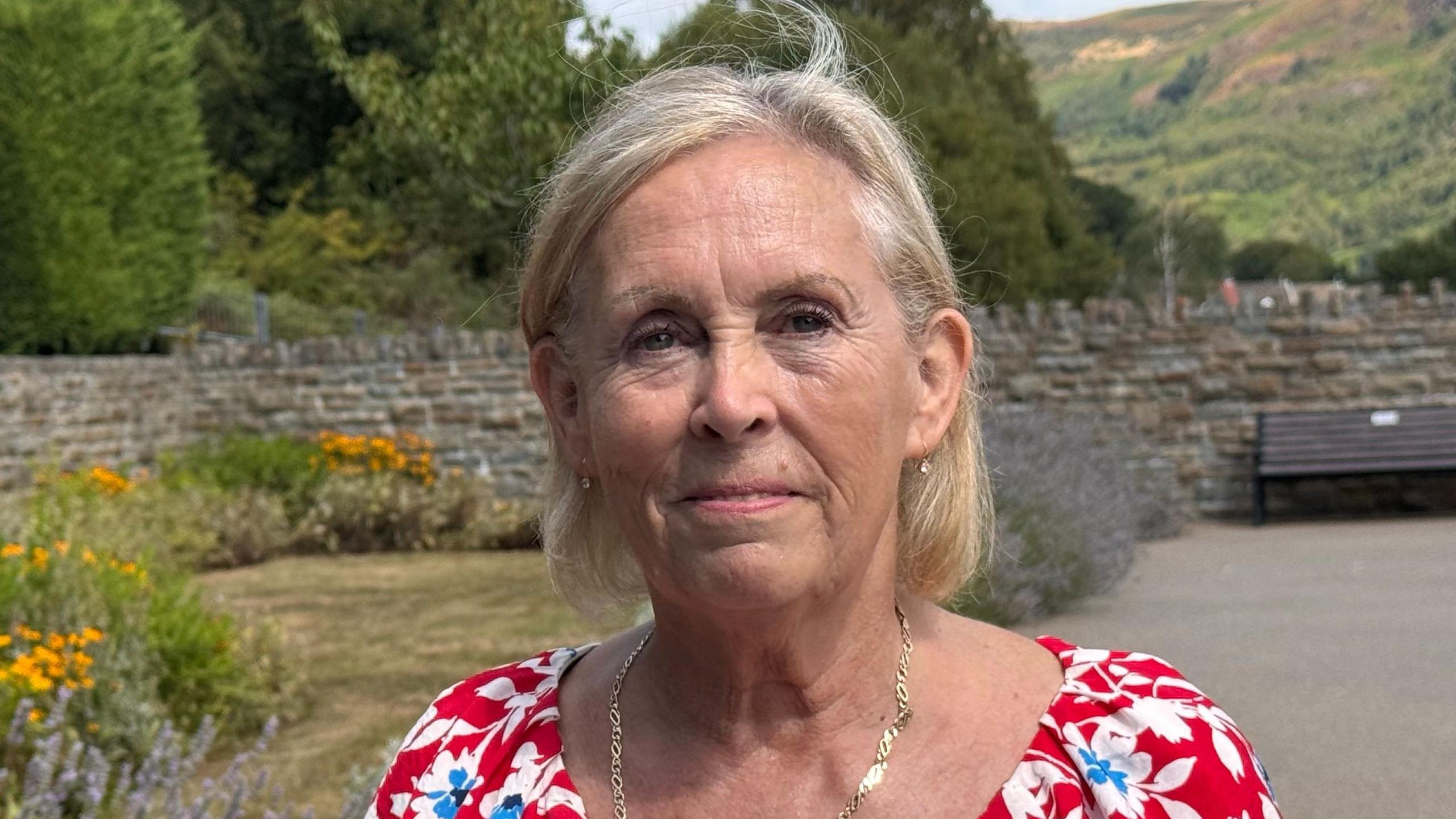 Janett wears a red floral dress and gold necklace. She has short light blonde hair and is smiling. It is a head and shoulders shot of her.