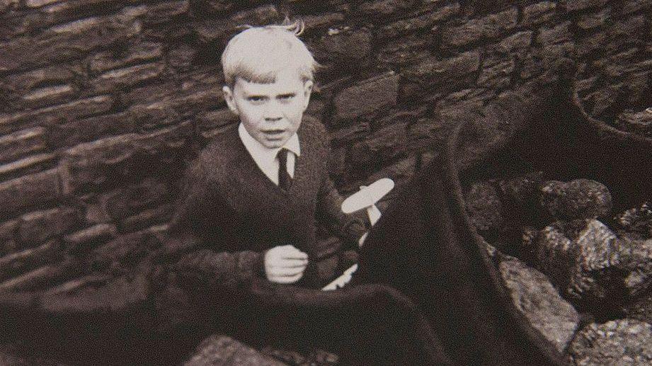 A young boy with white hair plays in the street behind sacks of coal