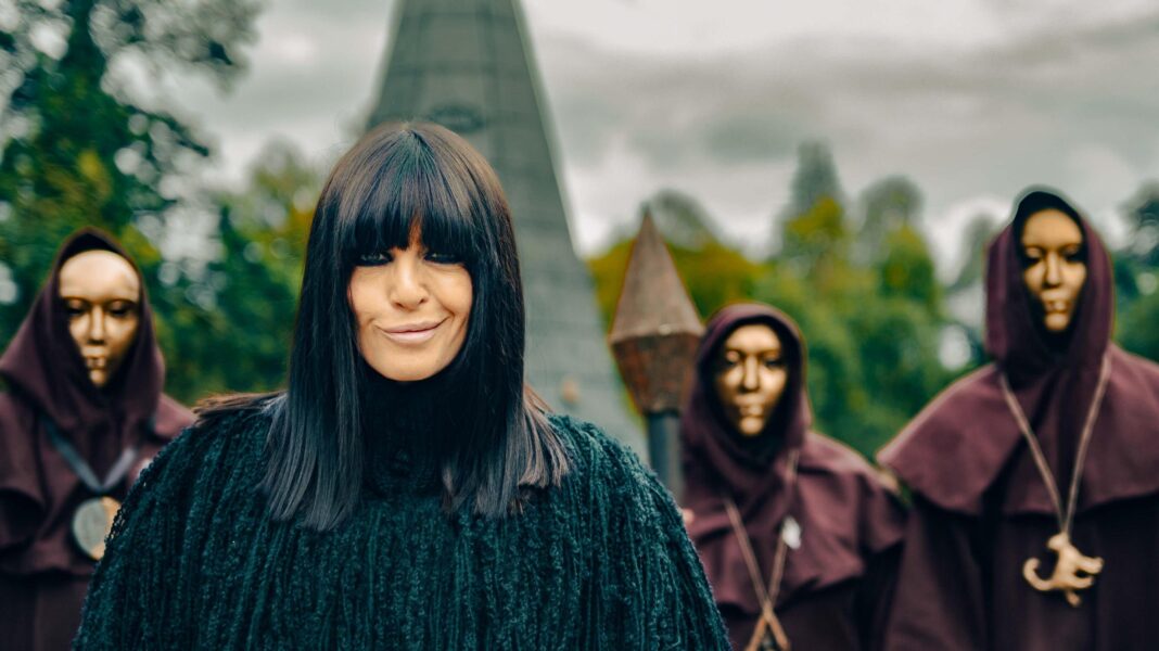 Celebrity Traitors 2 cast: Who are this year’s ‘big dogs’ and dark horses? Claudia Winkleman wears a black coat as she smiles at the camera. Behind her is part of a castle and three people in black hoods and golden masks.