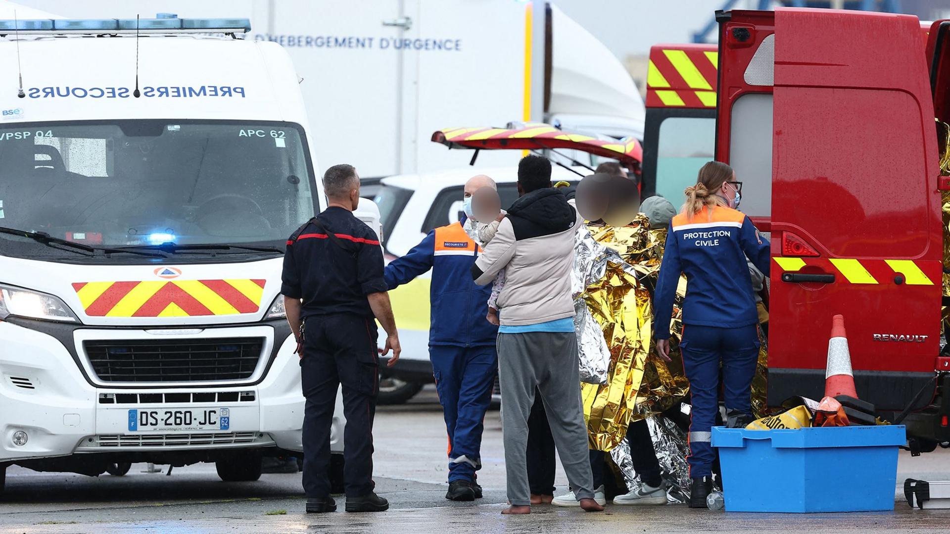 A group of people wearing tracksuits and coats collect gold survival blankets while they talk to staff wearing blue and orange uniforms. They stand by two vans.