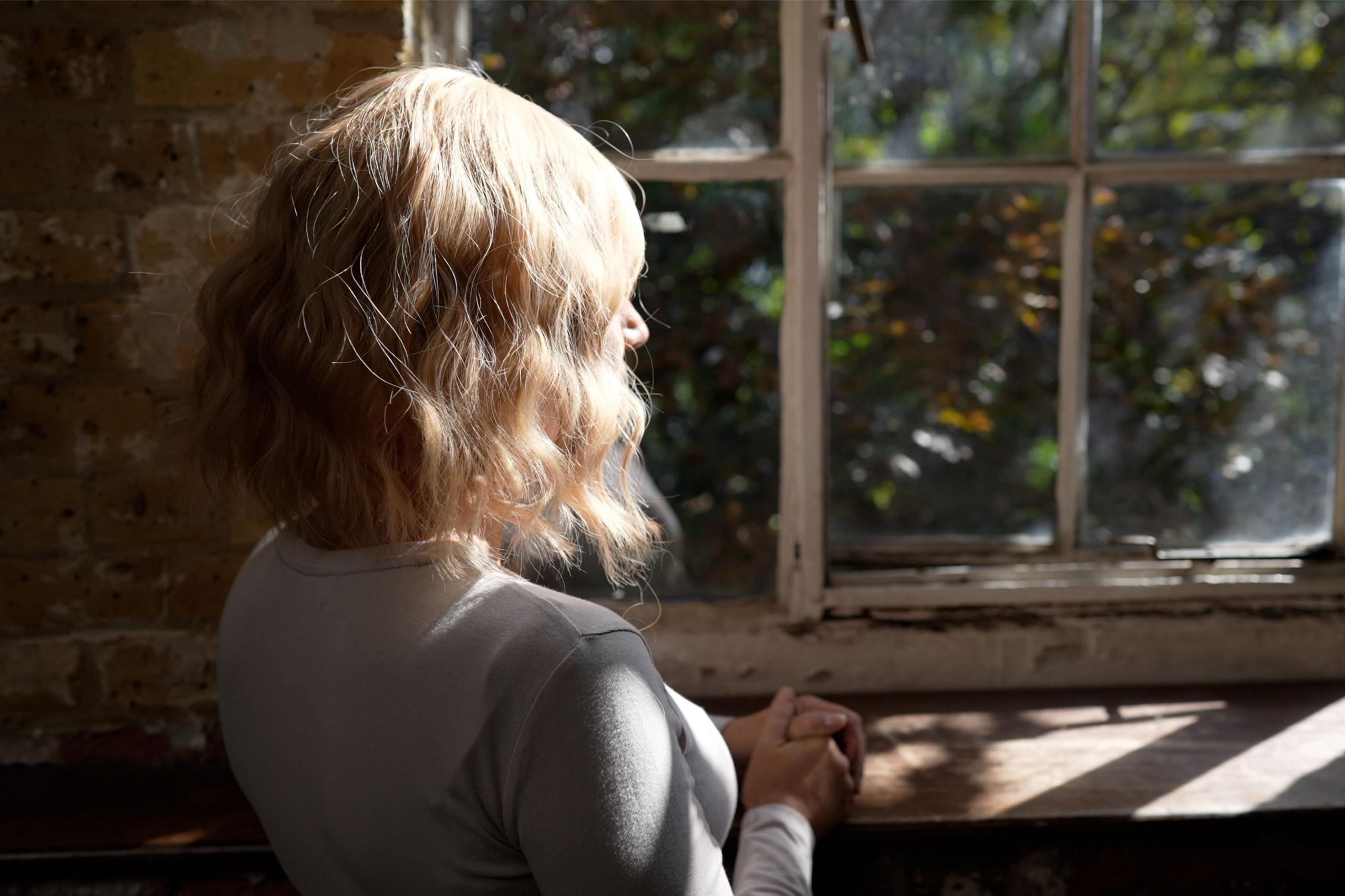 Laura sits by a sunlit window, her hands clasped as warm light catches her hair.