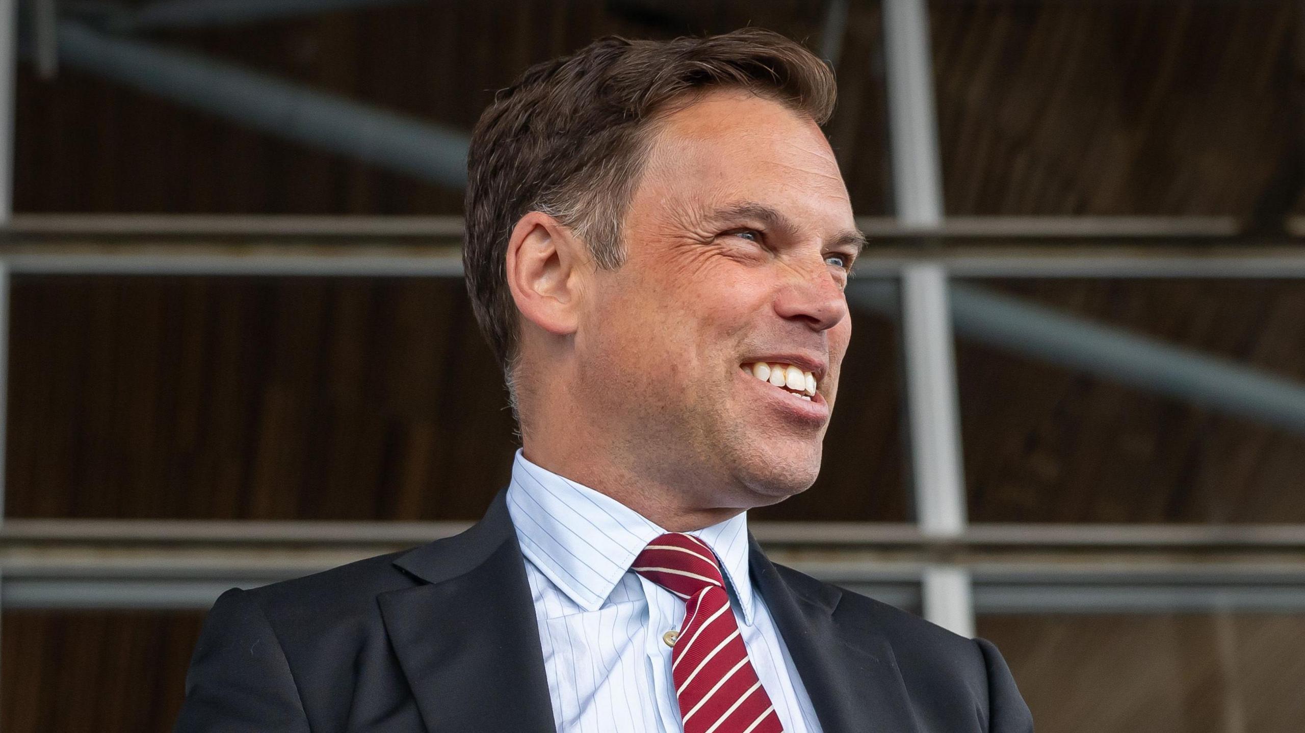 Ken Skates smiling off to the side of the image, wearing a dark suit and red and white tie, with a pin stripe shirt.