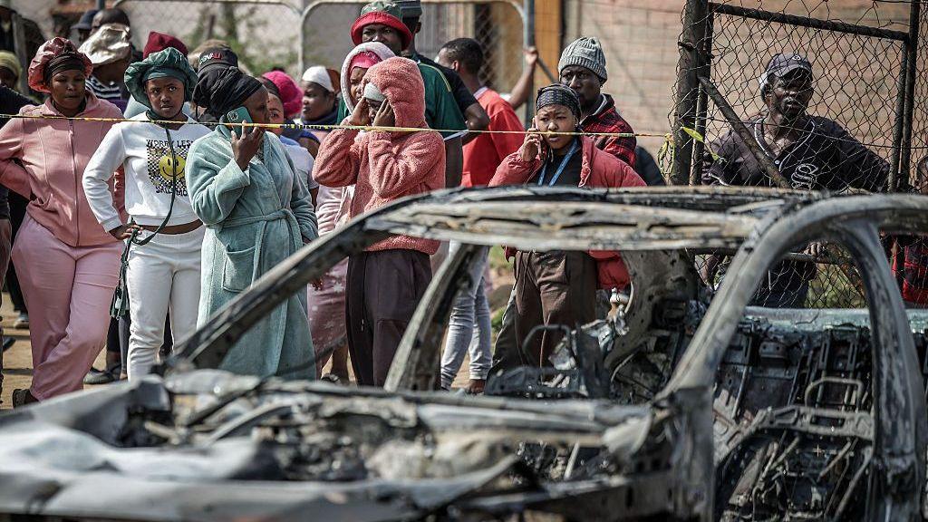 A crowd of people look on close to a burned out car