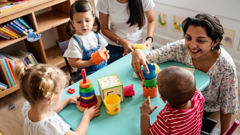 A group of very young children around a table, with a teacher. They have an assortment of toys. A bookshelf can be seen in the background.