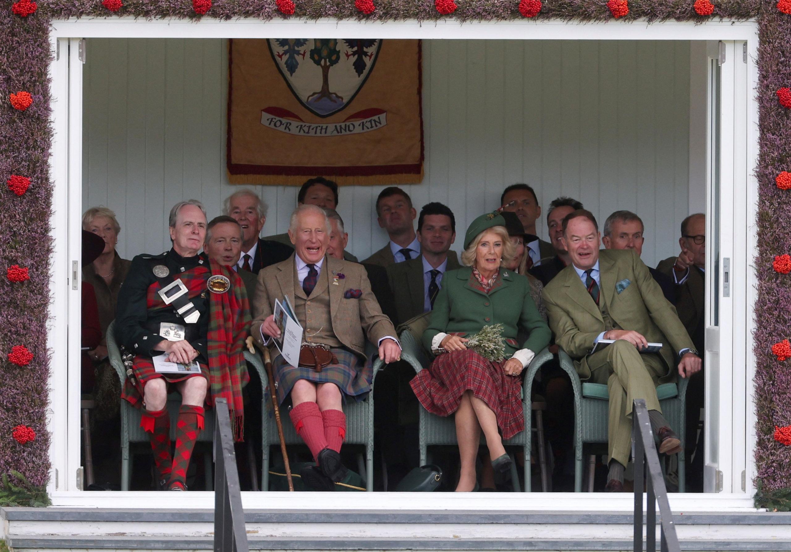 King Charles and Queen Camilla sit in a box watching an outdoor event - Charles is laughing