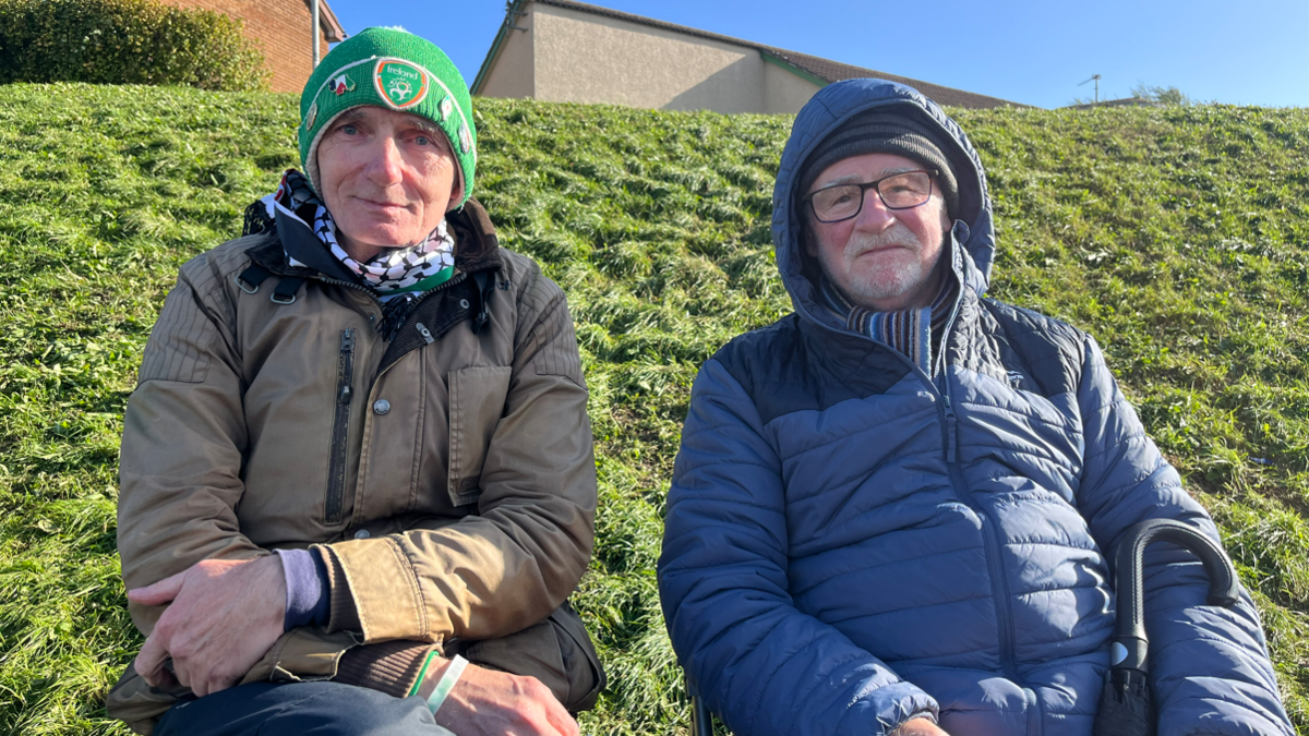Two men are sitting outside. One is wearing a green hat and has a brown coat on. The other man has a navy hat on and his hood is up. A hedge is behind them. The sky is clear.