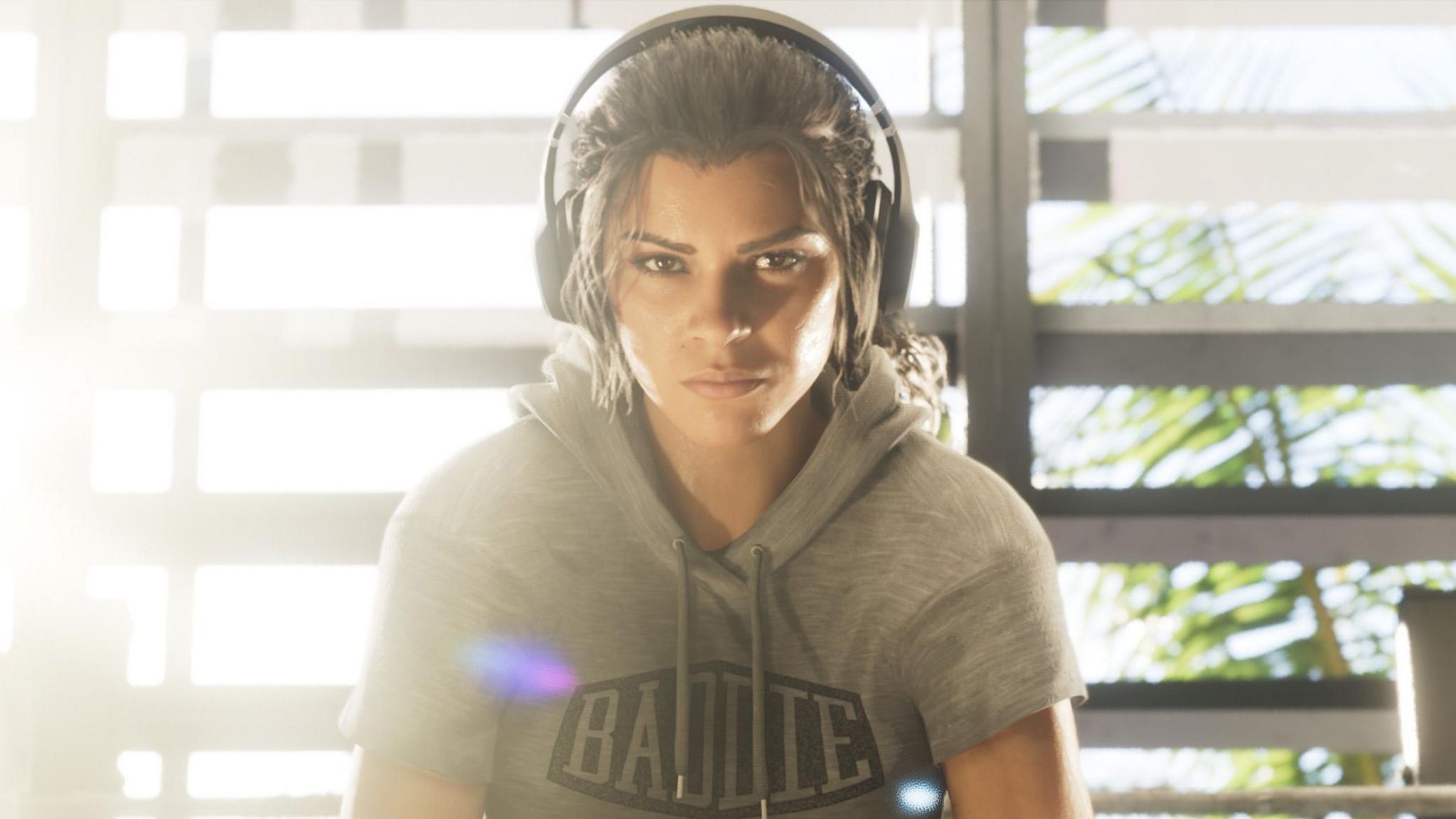 Computer generated but extremely lifelike image shows character Lucia in a gym, wearing large over-ear headphones and a grey hoodie with a black "Baddie" logo print. She's heavily backlit by the sun as she stares moodily at the viewer.