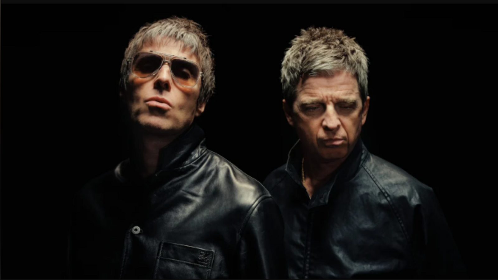 Liam and Noel Gallagher, both wearing black and looking serious in front of a black backdrop. Liam is wearing sunglasses