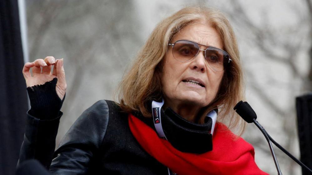 Gloria Steinem speaks at a lectern in Washington, wearing a red scarf and black jacket