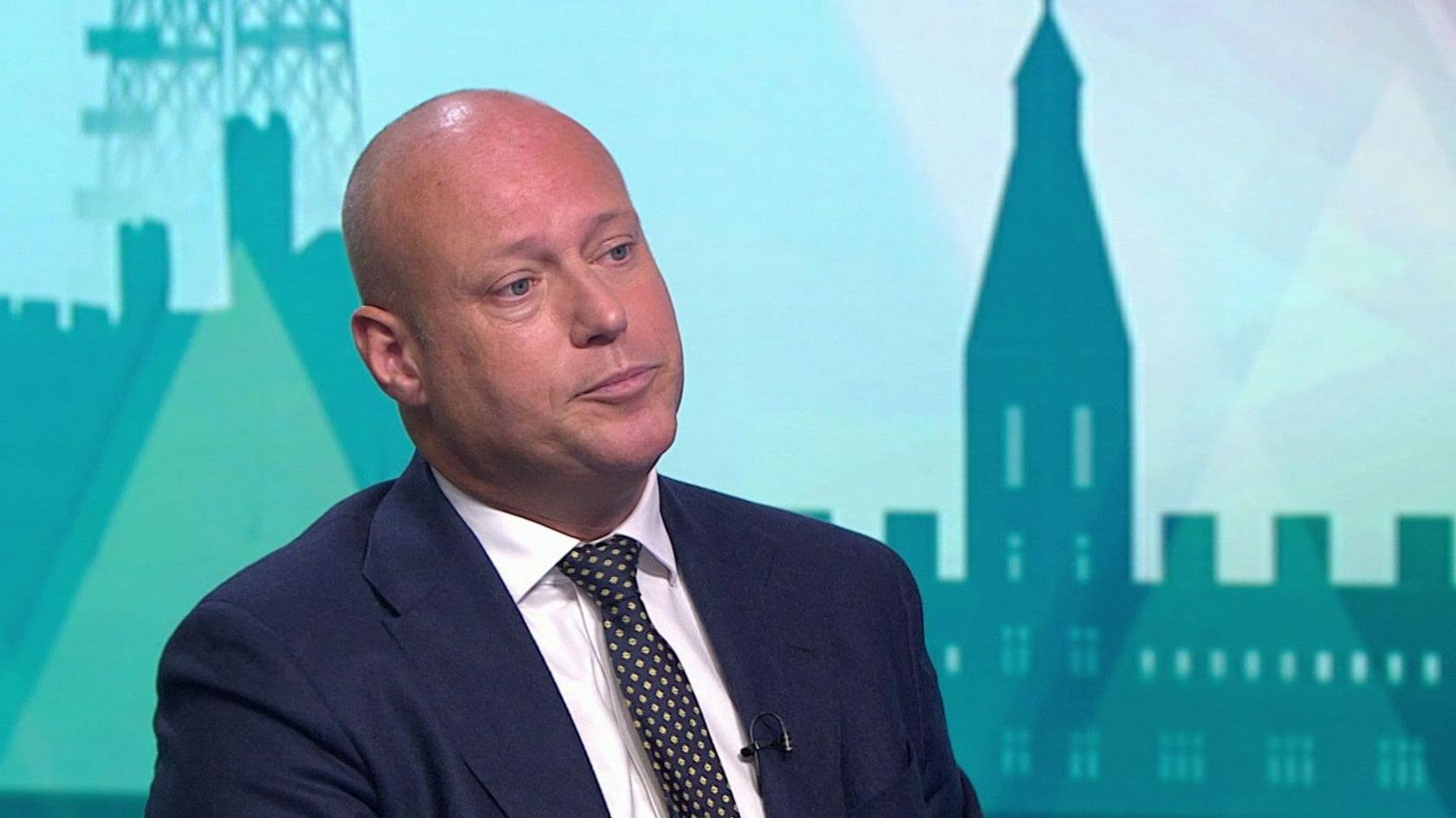 Dan Thomas being interviewed on Politics Wales . He is wearing a dark navy suit jacket, a white shirt and a dark tie with a light-coloured dot pattern. He has a bald head.