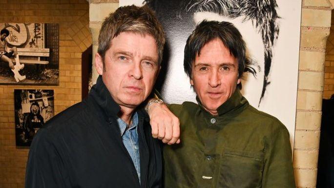 Noel Gallagher with Johnny Marr. It is a head and shoulders shot of the two of them. Noel is on the left and has short grey hair, and a dark blue jacket and a denim shirt underneath. Marr has a khaki green jacket buttoned up and short black hair.