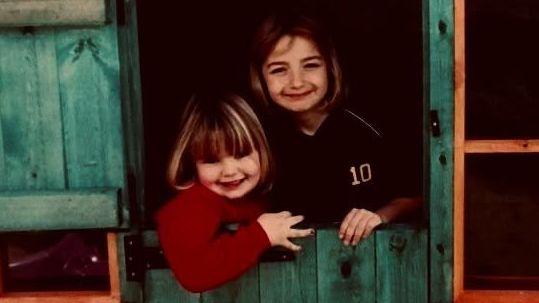 Maddy Cusack with her younger sister Olivia as children. They smile at the camera, leaning over a stable door, Olivia leaning into her older sister who is taller than her.