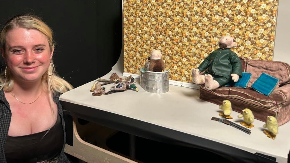 Stop-motion student Annie Buckeridge stands beside her small film set, which includes a fabric farmer dressed in a green coat and sitting on a tiny sofa next to three small yellow model chicks. Annie has long blonde hair, wears a dark top and is smiling at the camera.