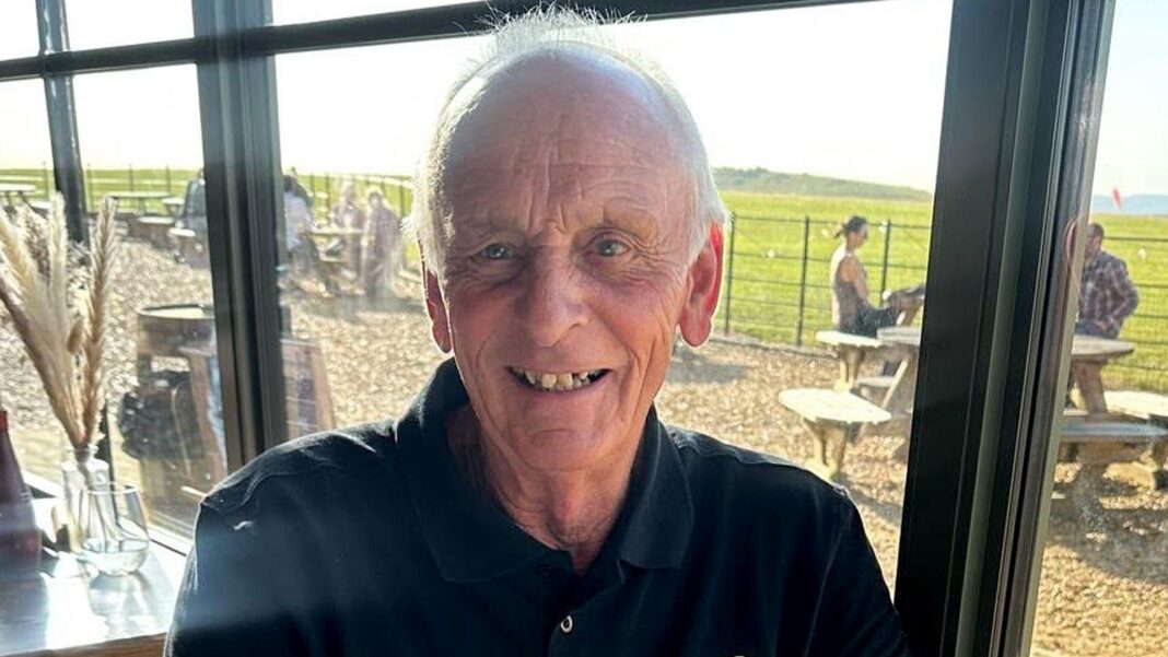 ‘My cancer spread while I waited months for surgery’: The cost of England’s cancer wait times An older man wearing a black polo shirt, sits at a restaurant table with views of the countryside. He is smiling at the camera.