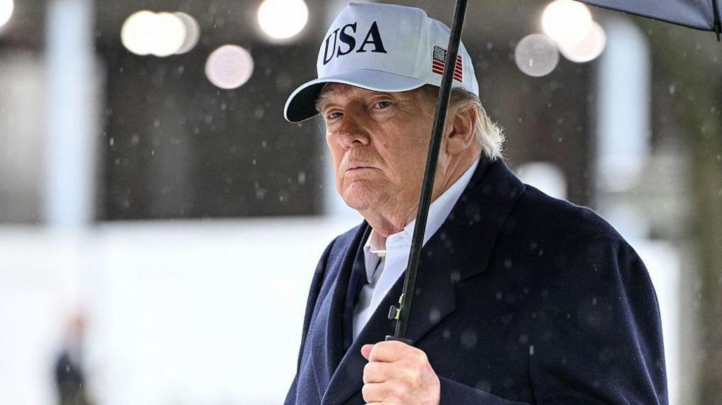 President Trump holds an umbrella as rain drops fall around him. He wears a dark suit and a white baseball cap with USA written on the front and an American flag on the side.