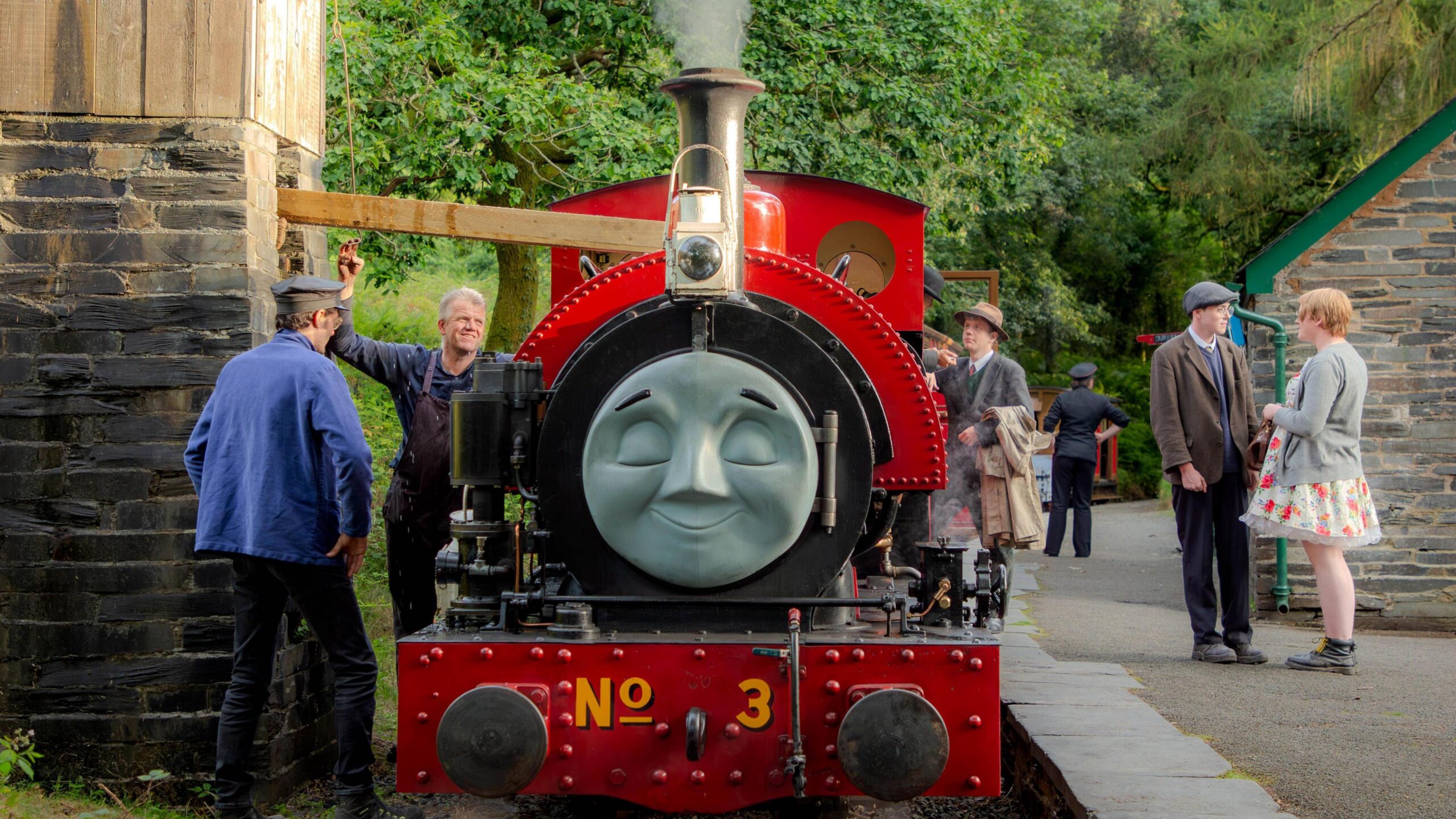 A small red steam locomotive stands in a countryside station next to a water tower. The locomotive has a face attached to the smokebox at the front, making it look like one of the characters from Thomas the Tank Engine. People in 1950s period costume stand on the platform