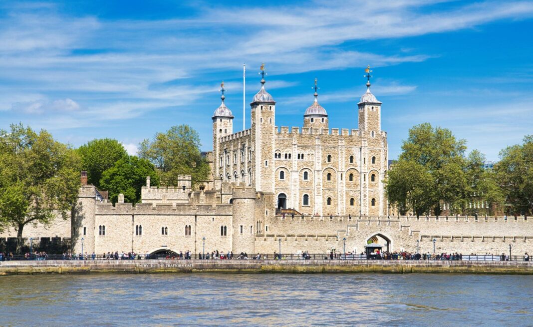 The Tower of London viewed from across the River Thames, showing its stone walls, corner towers with domed roofs, and surrounding grounds under a blue sky, with people walking along the riverside.