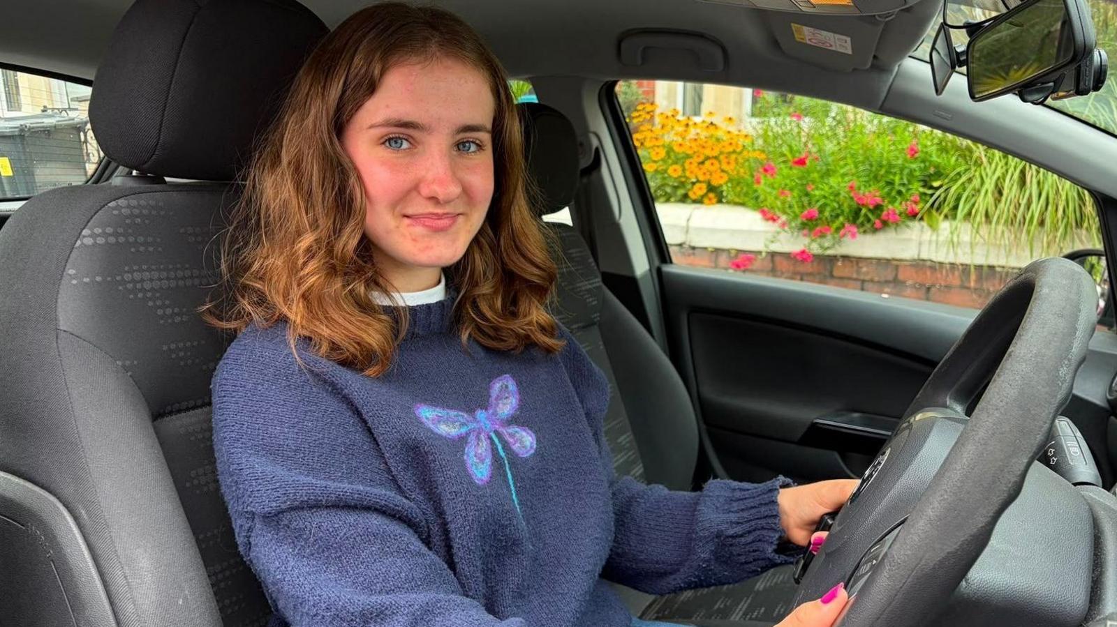 Megan sits in her car. she is smiling at the camera and has reddish to brown hair cut at her shoulders. She is wearing a blue jumper with a dragonfly on.