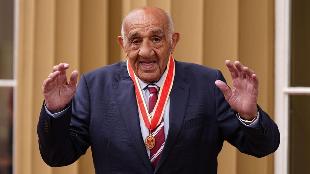 Wigan prepares to say goodbye to Sir Billy Boston Sir Billy Boston is wearing a navy suit and light blue shirt and red and white tie. He is wearing an insignia and holding both hands up and smiling at the camera.