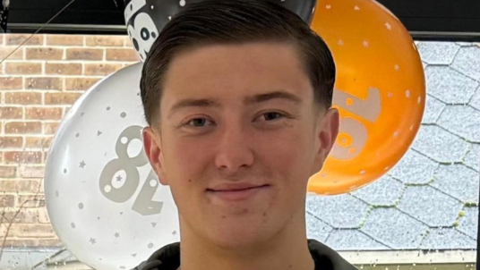 Henry Nowak stands smiling in front of orange, white and black balloons with "18" printed on them.