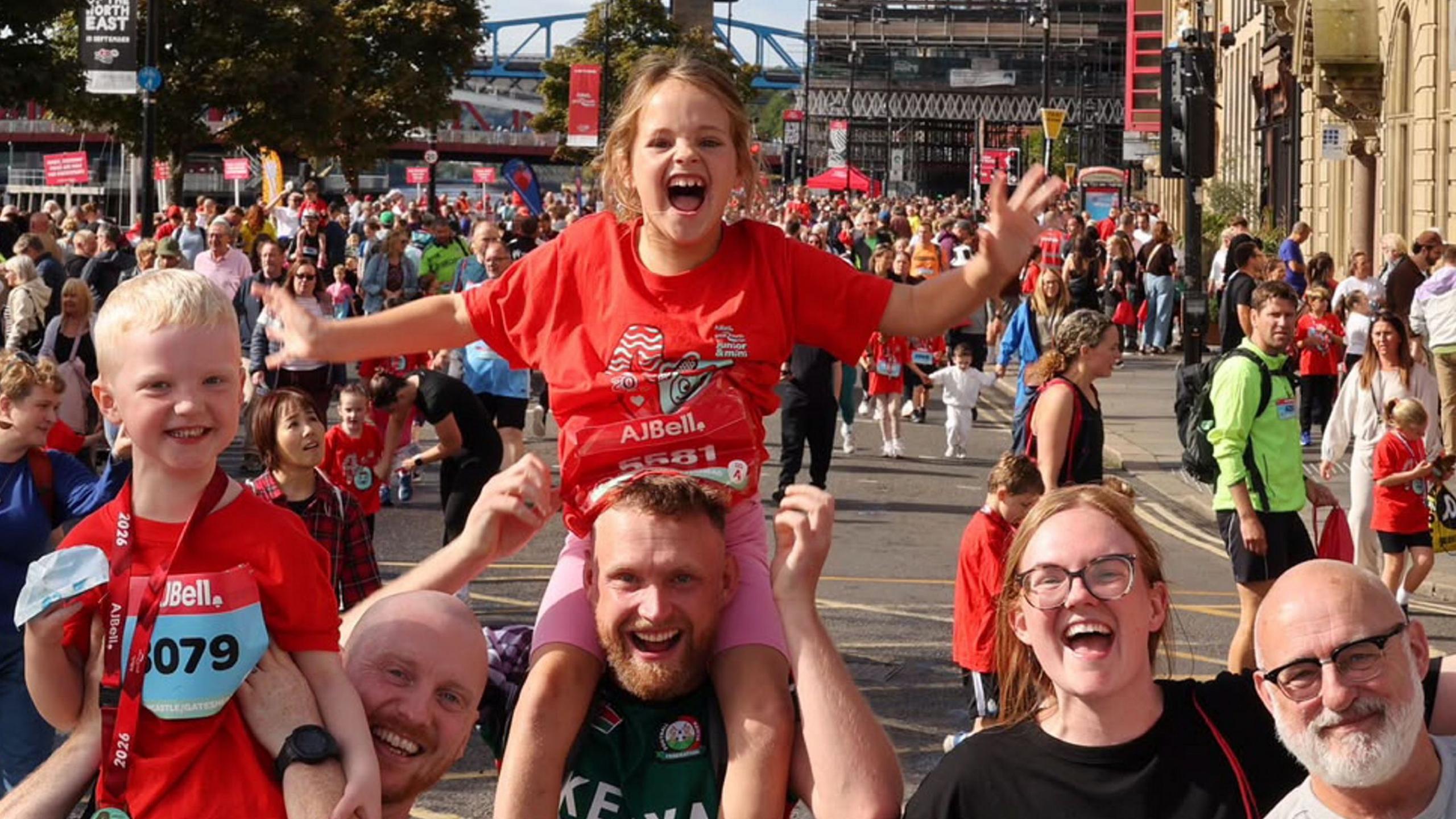 Two children are perched on the shoulders of a group of four adults. They are all smiling and cheering at the camera as they walk along the road which is full of crowds of people. The children are wearing red and blue running numbers.