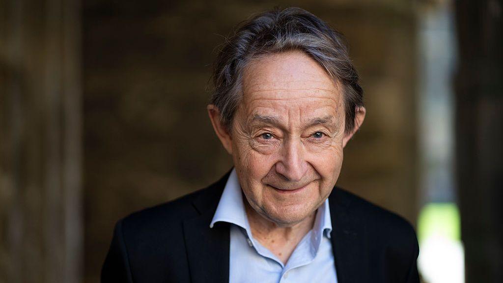 Photo of Sir Anthony Seldon in 2025. He is wearing a light-blue shirt and a black blazer and is smiling at the camera. The background behind him is blurred.