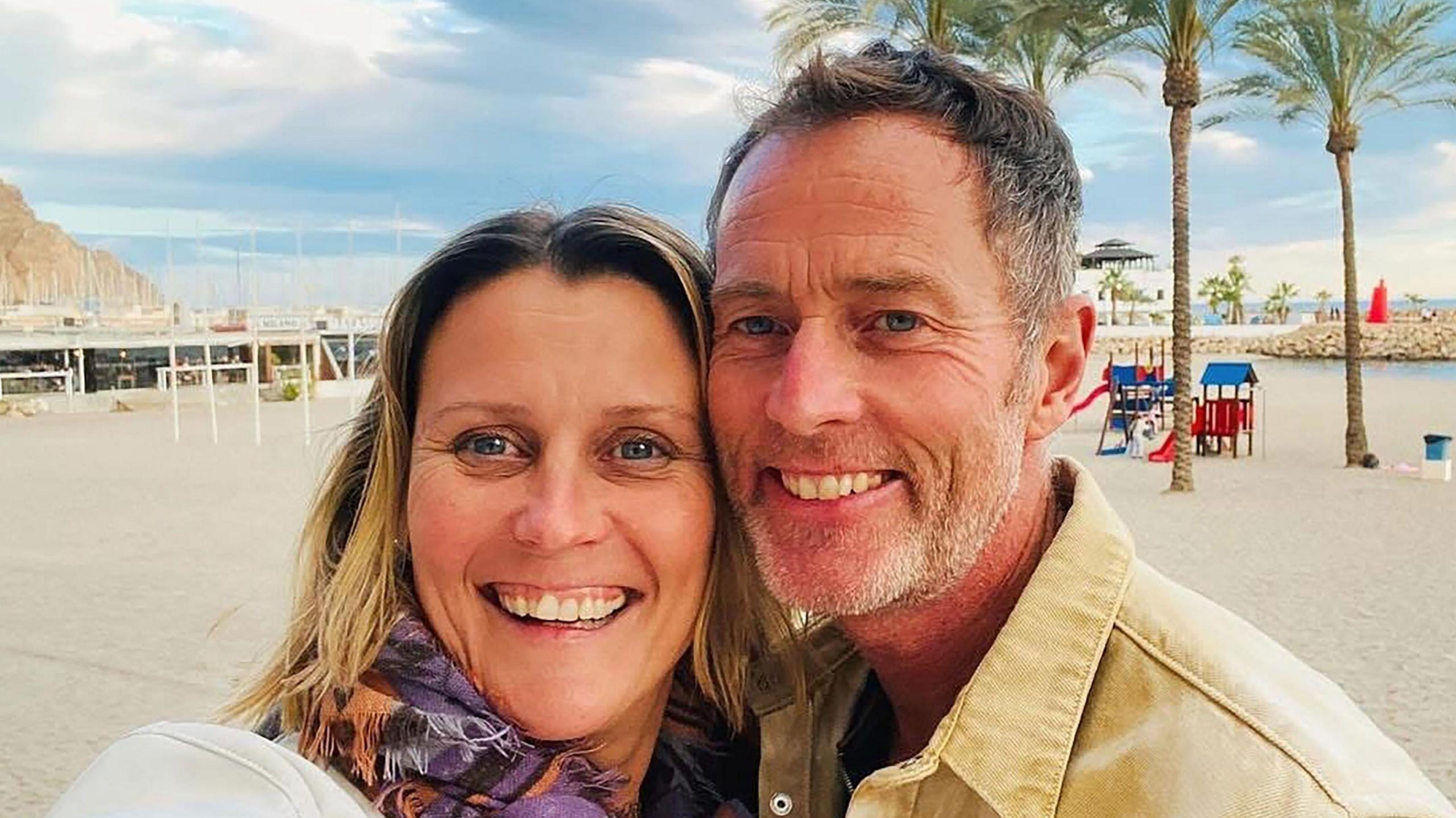 A couple on a beach. It is sunny and the sand is white. Behind them are some trees.