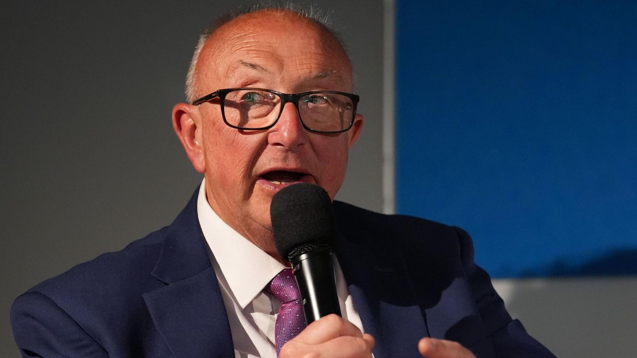 Colin Sutton is wearing black-rimmed glasses and a navy suit, white shirt and purple patterned tie. He is holding a microphone close to his mouth and looks to be in a corporate environment.