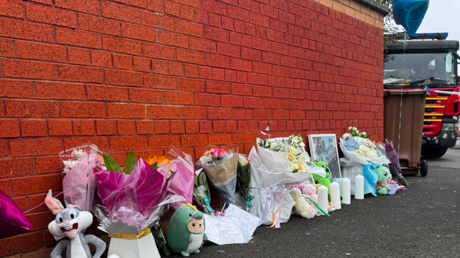 Image shows a number of floral tributes and cuddly toys left along a wall, with a fire engine in the background.