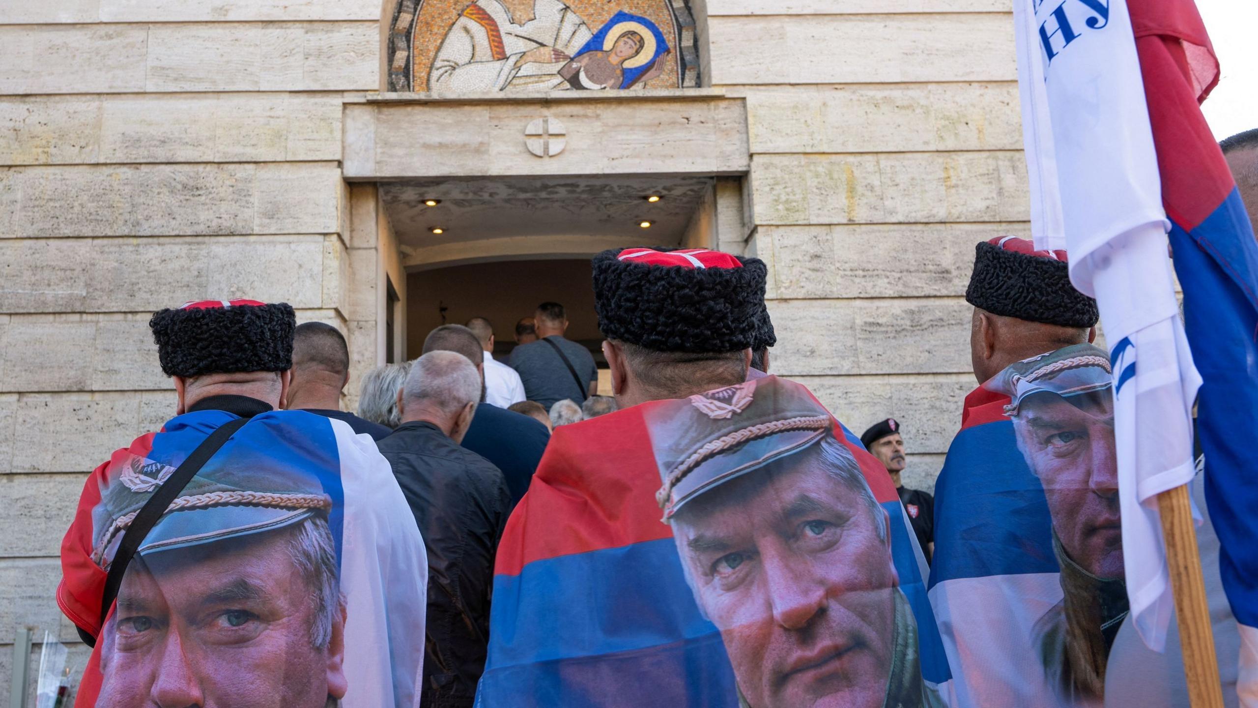 Men wearing flags as capes, with a picture of former Bosnian Serb wartime general Ratko Mladic, stand amongst people gathered at the Church of St. Luke, where the body of Mladic lies in state before burial, in Belgrade, Serbia, September 7, 2026.