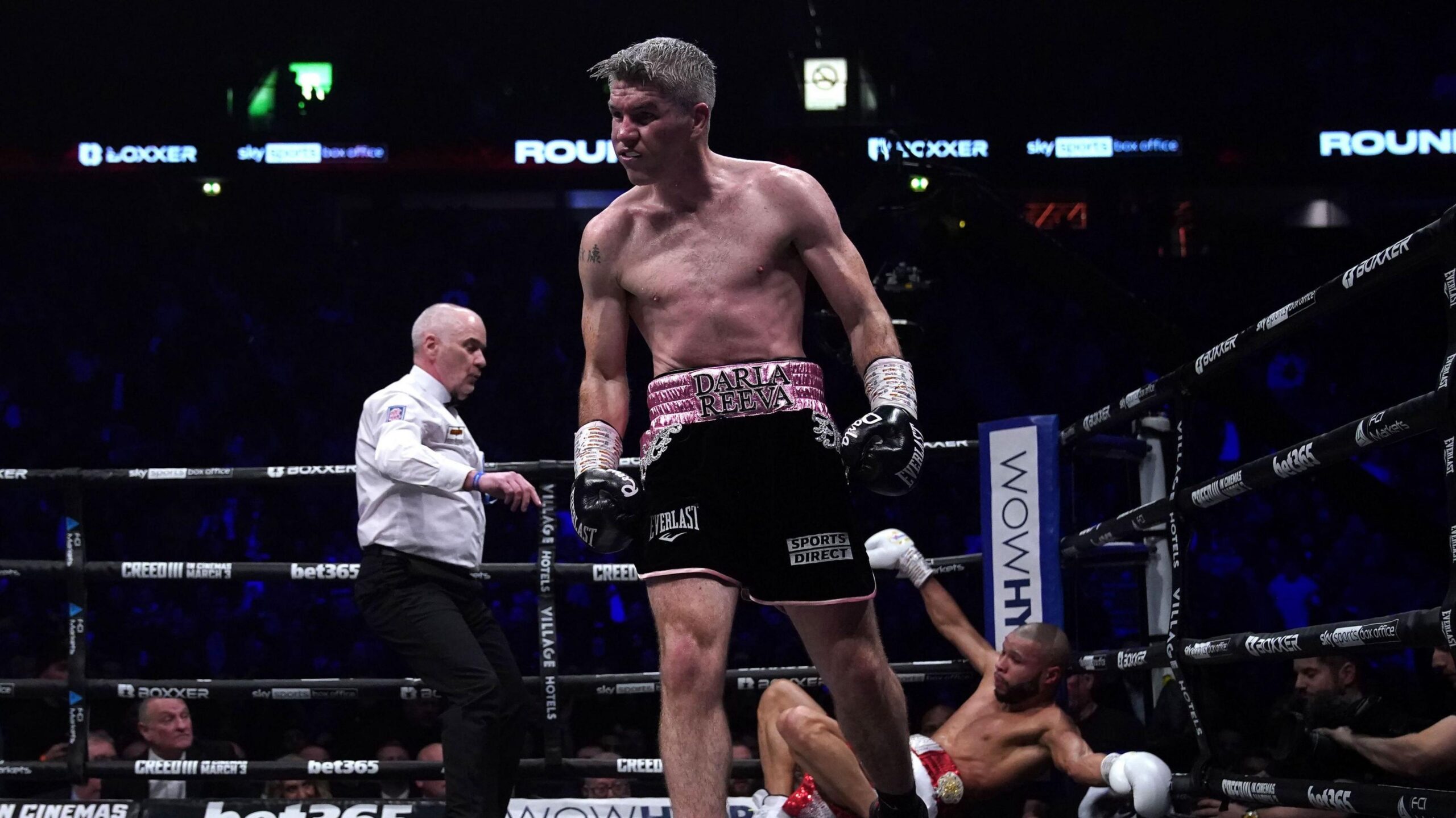 A boxer, wearing black shorts and with grey hair, turns away after knocking down his opponent, who is black and has a shaved head, and is on the canvas leaning against the corner of the ring. A referee in a white shirt has his arm outstretched.
