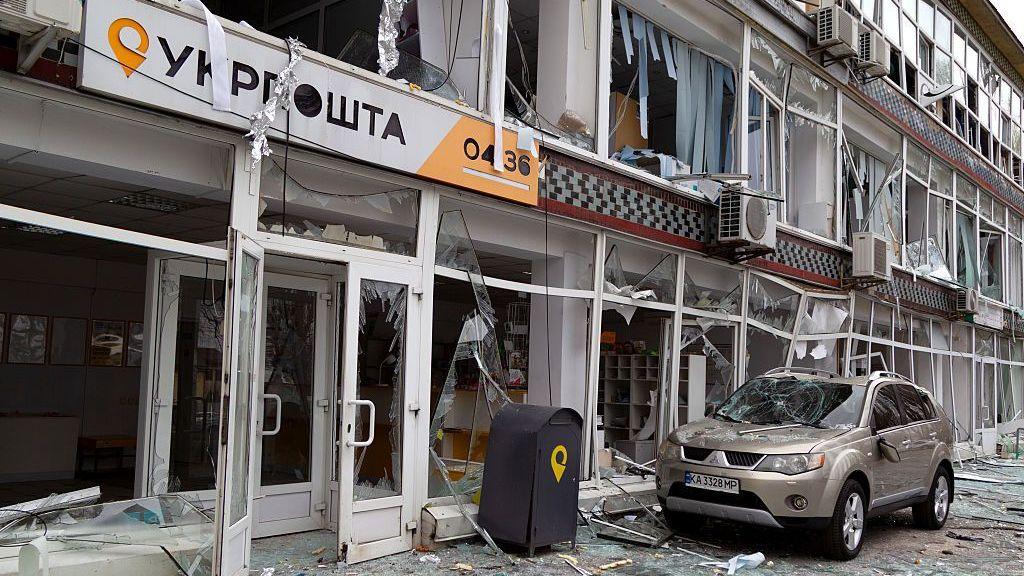 A shopfront, with a sign reading 'Ukrposhta' in Ukrainian, is seen heavily damaged with the windows blown out and glass shattered on the pavement outside. A car, which is parked outside, has a smashed windscreen.