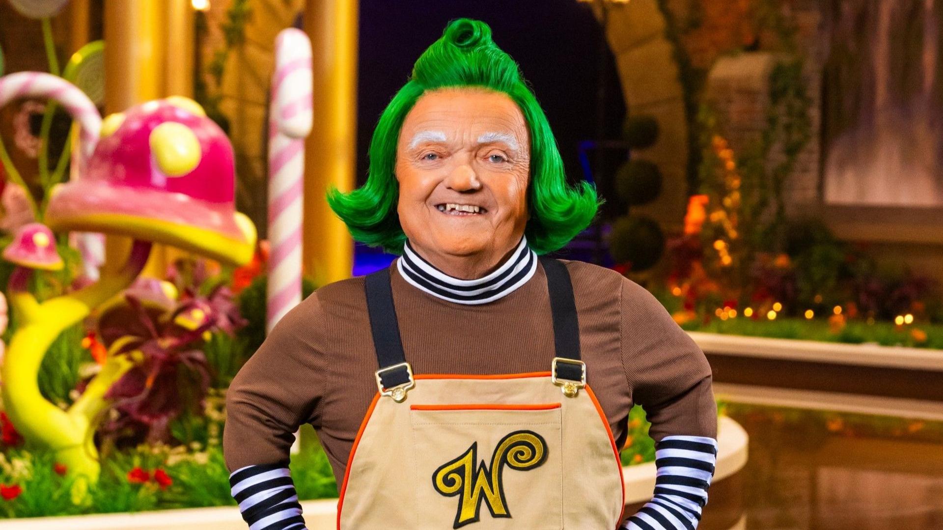 A recent photo of the actor Rusty Goffe dressed as an Oompa Loompa on set in Willy Wonka's factory with tall candy canes and a chocolate river behind him