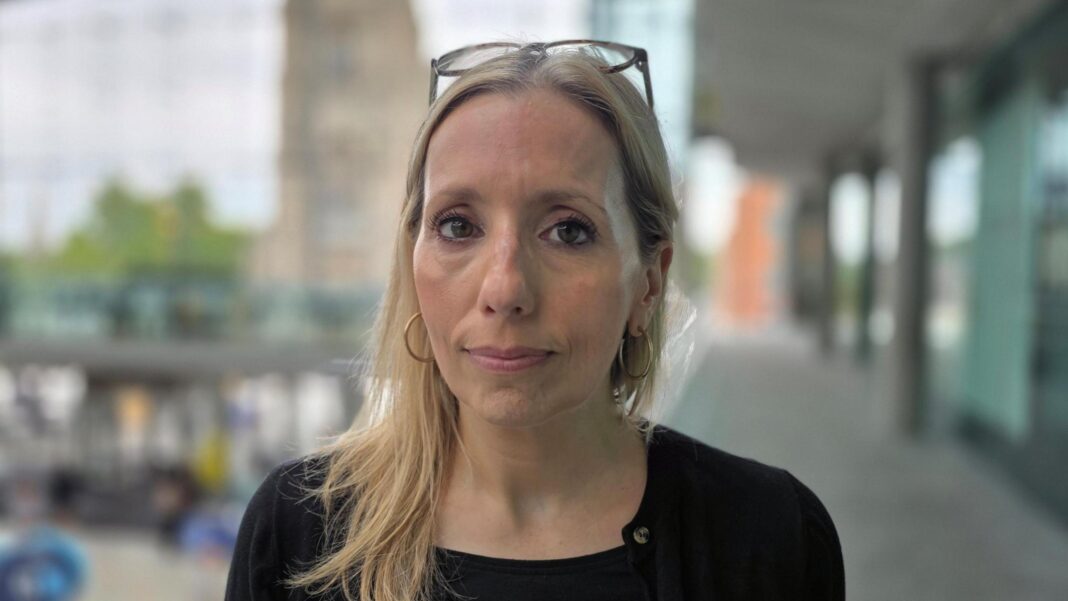 Police commissioner may cancel tour after backlash Sarah Taylor is wearing a black top. Her glasses are placed on top of her head. She is standing inside Norwich's Forum building, and a church tower is in the very background the other side of the glass frontage, out of focus.