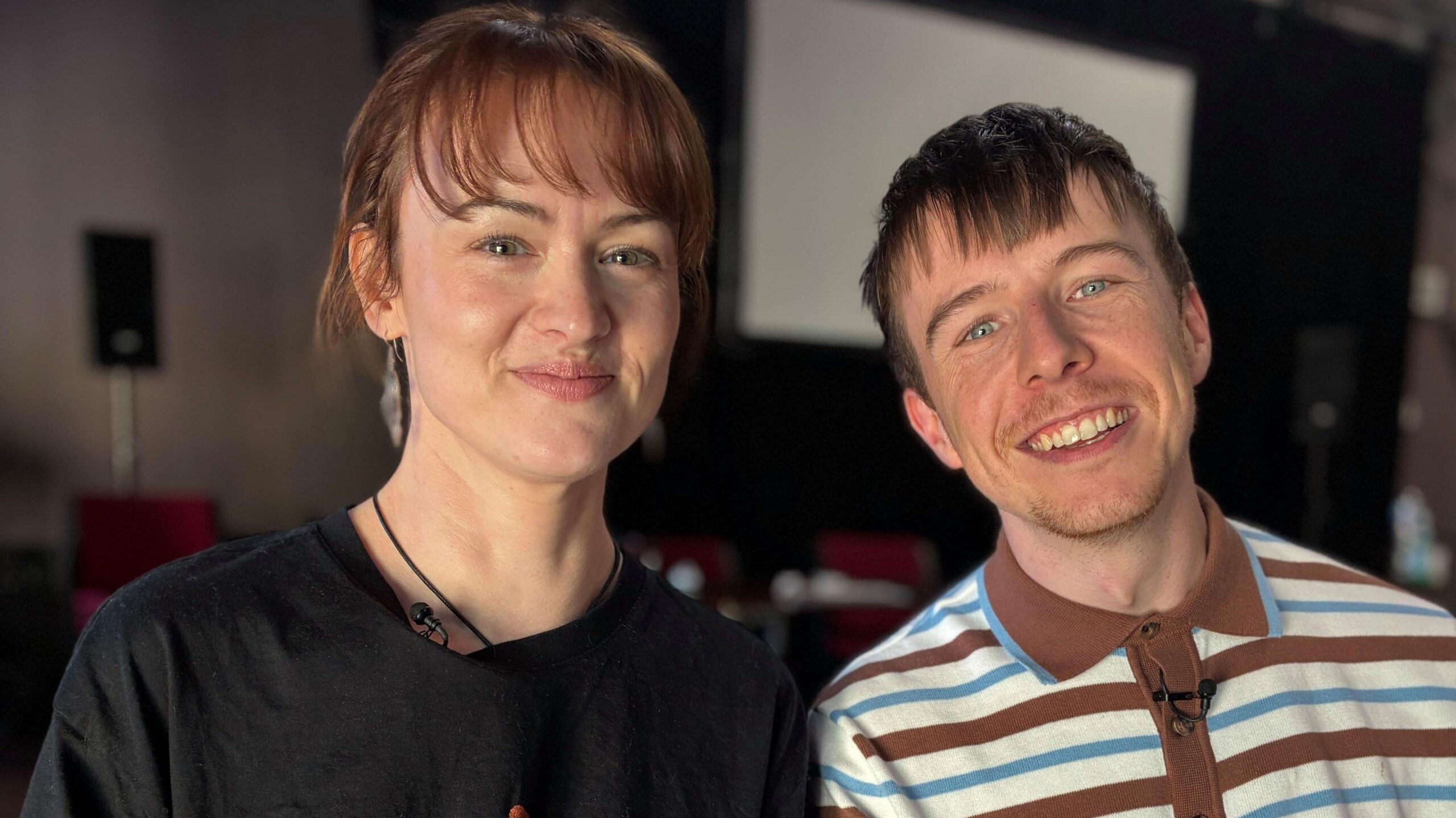 Sam Neale has red hair and is smiling and has a black t-shirt on. Ryan Nolan is smiling with his mouth open, and has a buttoned-up brown, white and blue striped shirt on. He has stubble and short brown hair.