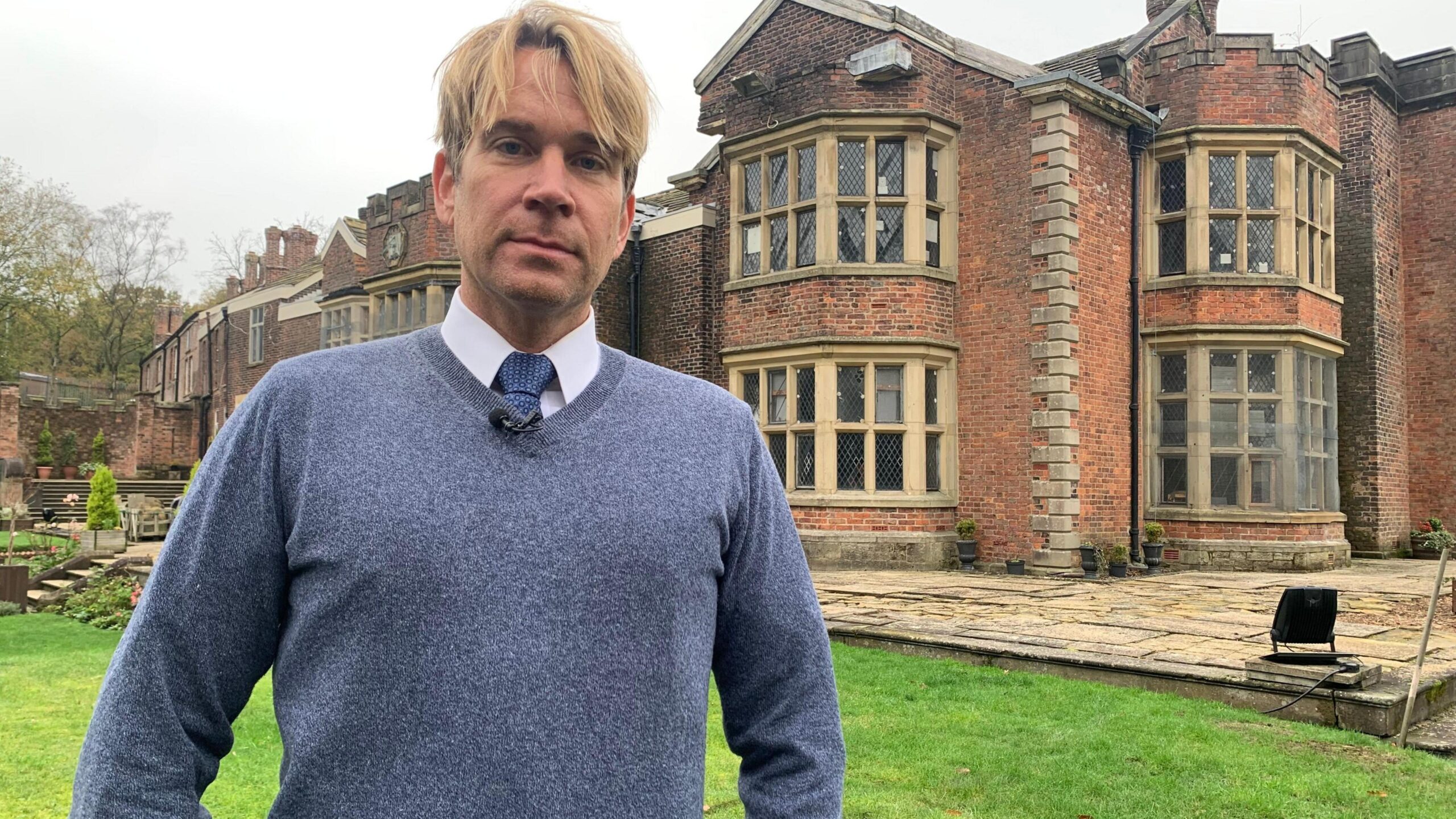 Hopwood DePree stands outside Hopwood Hall, in Middleton, Rochdale. He has blond hair and his wearing a tie under a blue jumper.