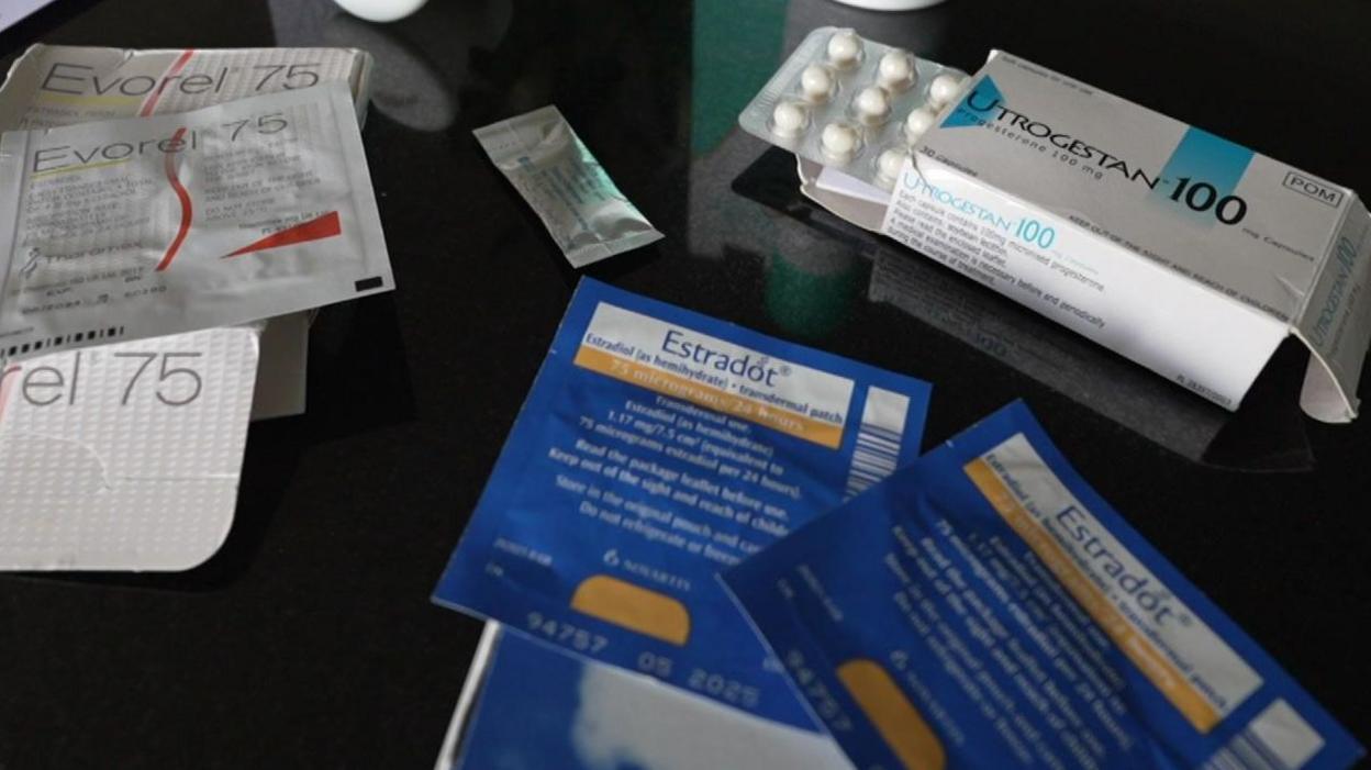 Different types of HRT are laid out on a table, including tablets, patches, spray and gel. In the centre is a small sachet of testosterone gel.