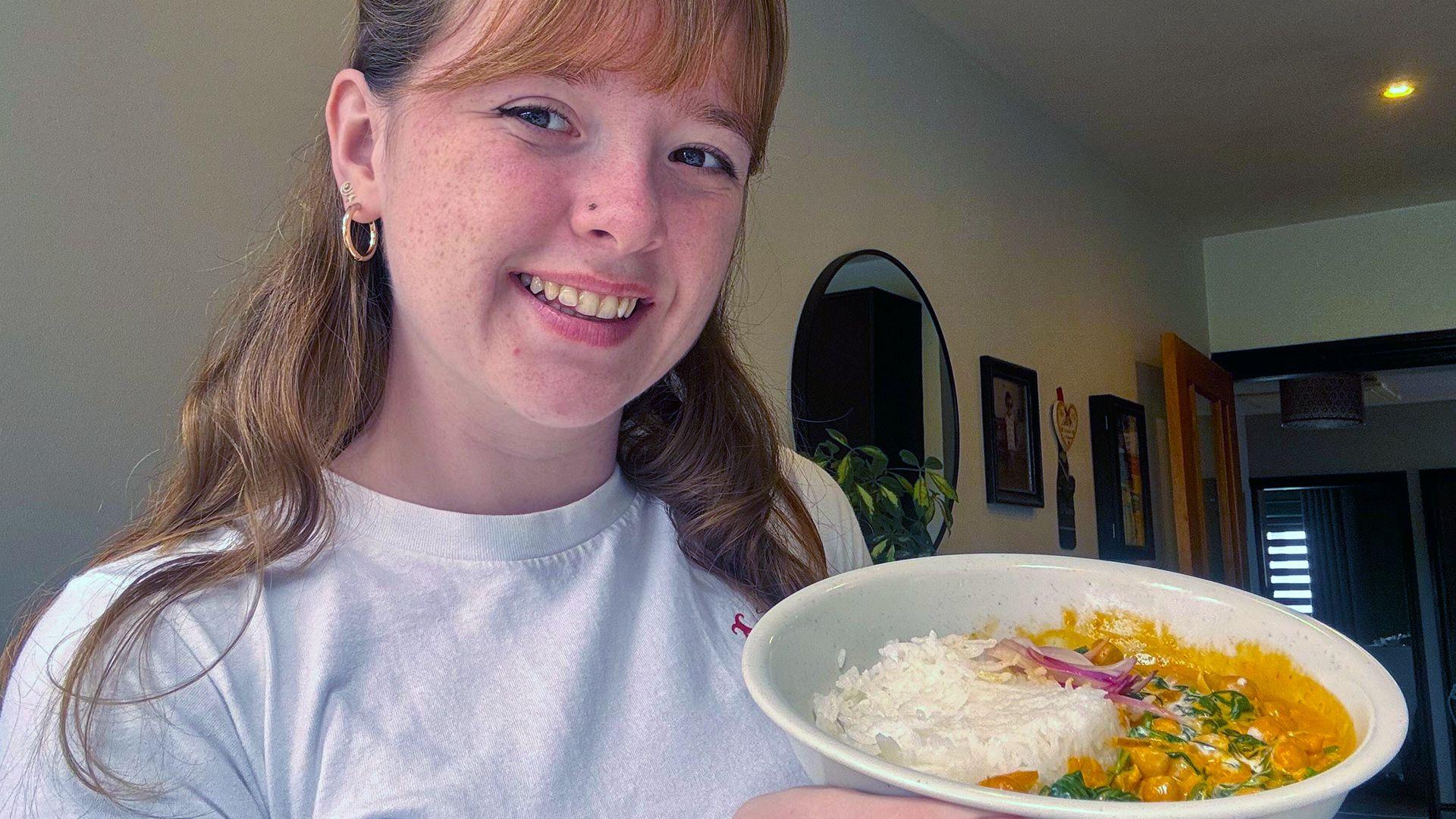 Mia with light brown hair with a fringe wearing a white t shirt is smiling holding a bowl of chickpea curry.
