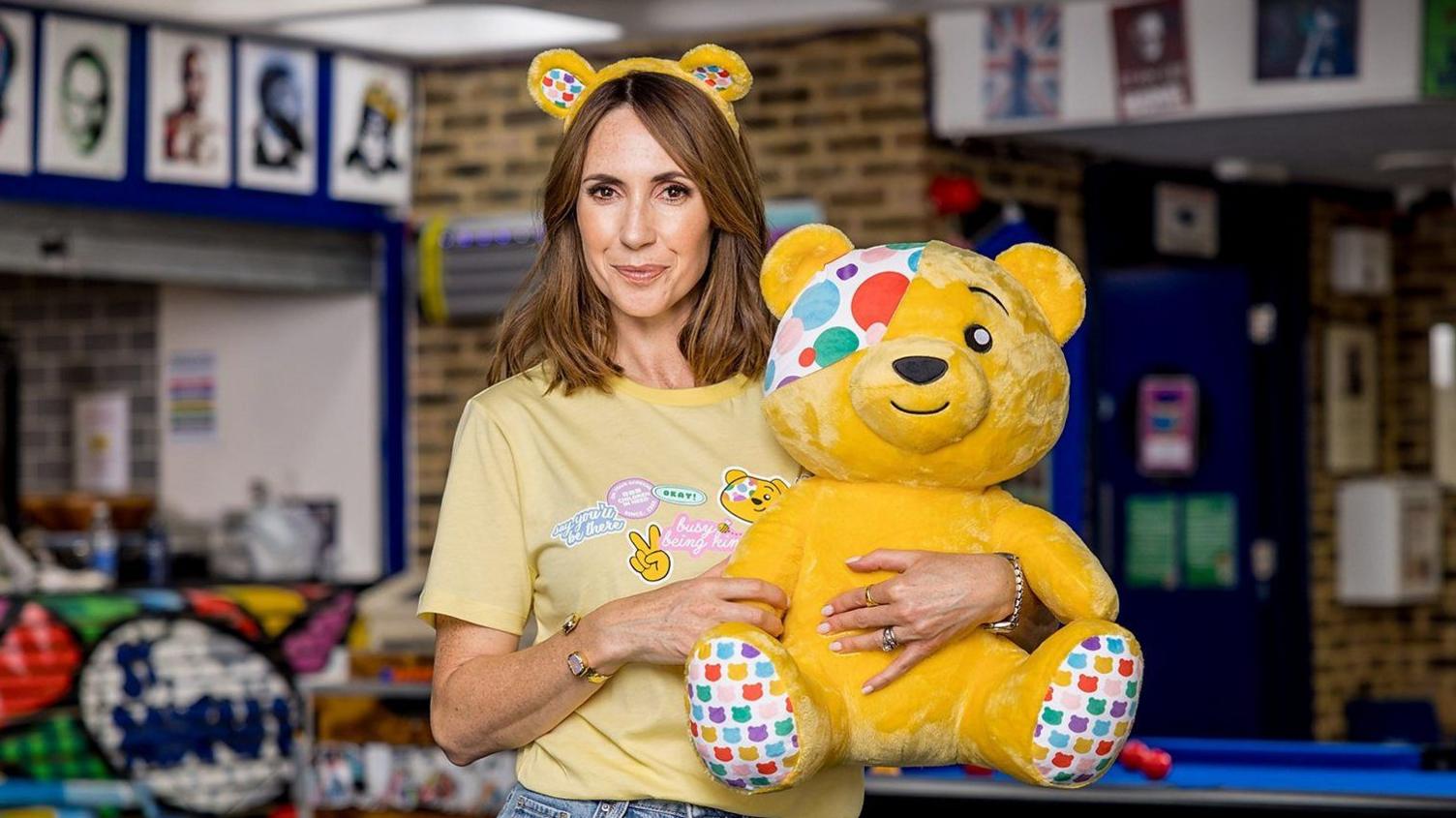 One Show host Alex Jones is wearing a yellow T-shirt, a pair of false ears and holding a cuddly toy