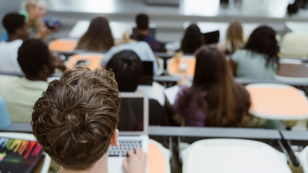 ‘Super-university’ created amid funding squeeze A group of students sits in a tiered lecture hall, listening to a presentation at the front of the room. In the foreground, one male student with brown hair types on a laptop.