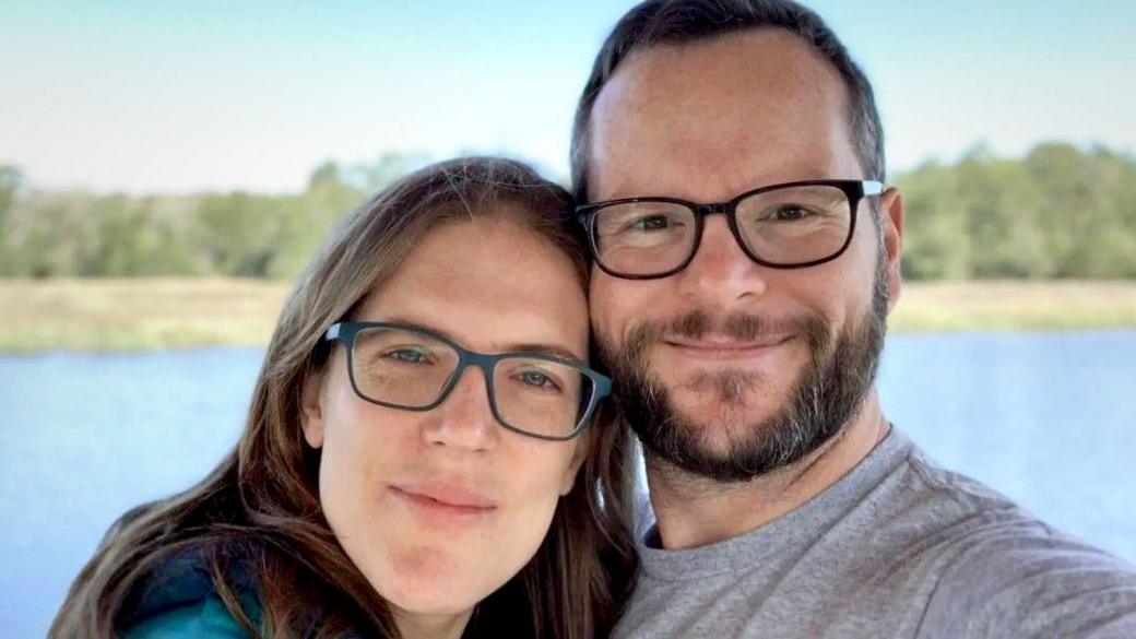 Katie and Alan Donegan smile at the camera while both wearing glasses during a selfie in front of a lake and trees against a blue sky.