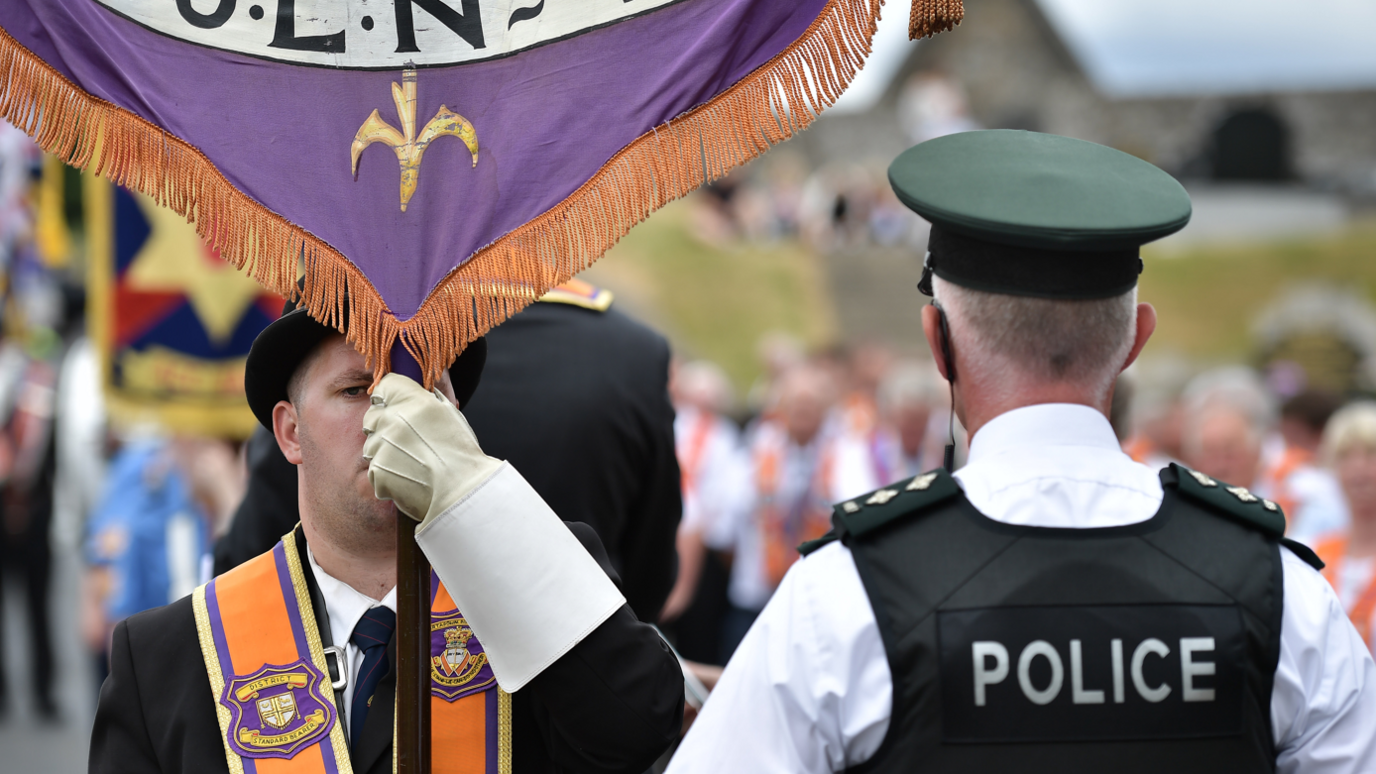 A man is wearing an orange sash and a black bowler hat and holding a banner. A policeman has his back to the camera. He has a black vest on stating police in white lettering. He has a cap and a white shirt on. A parade is out of focus in the background.