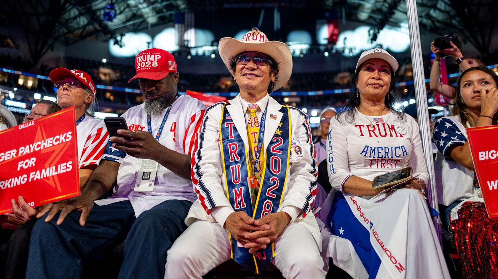 A man in a white suit with Trump 2028 written on a scarf and a cowboy hat sits and smiles at the Trump midterm convention